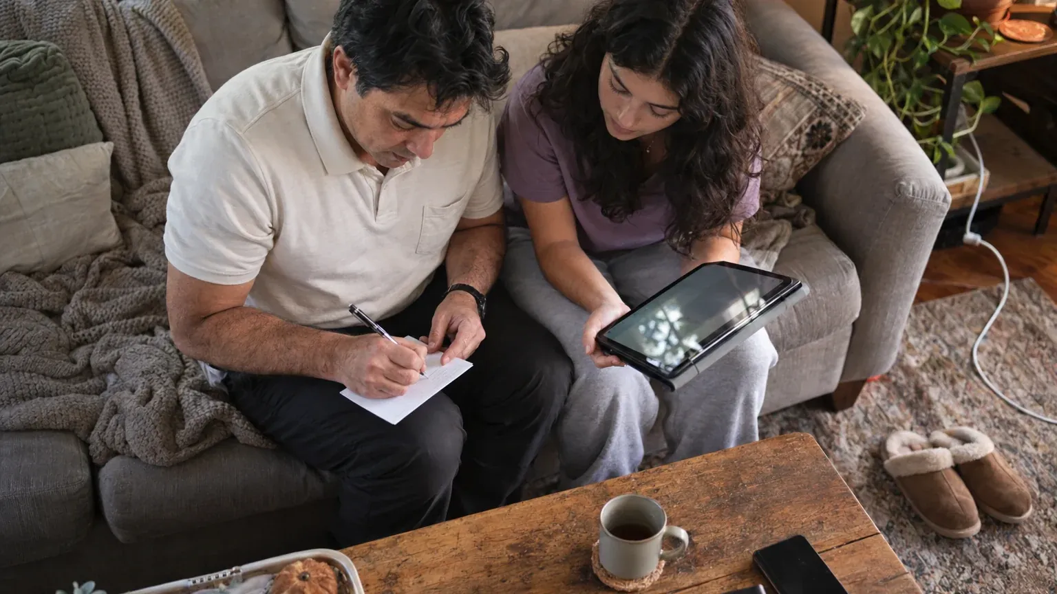 A man takes notes on paper while sitting on a sofa next to a woman with a tablet